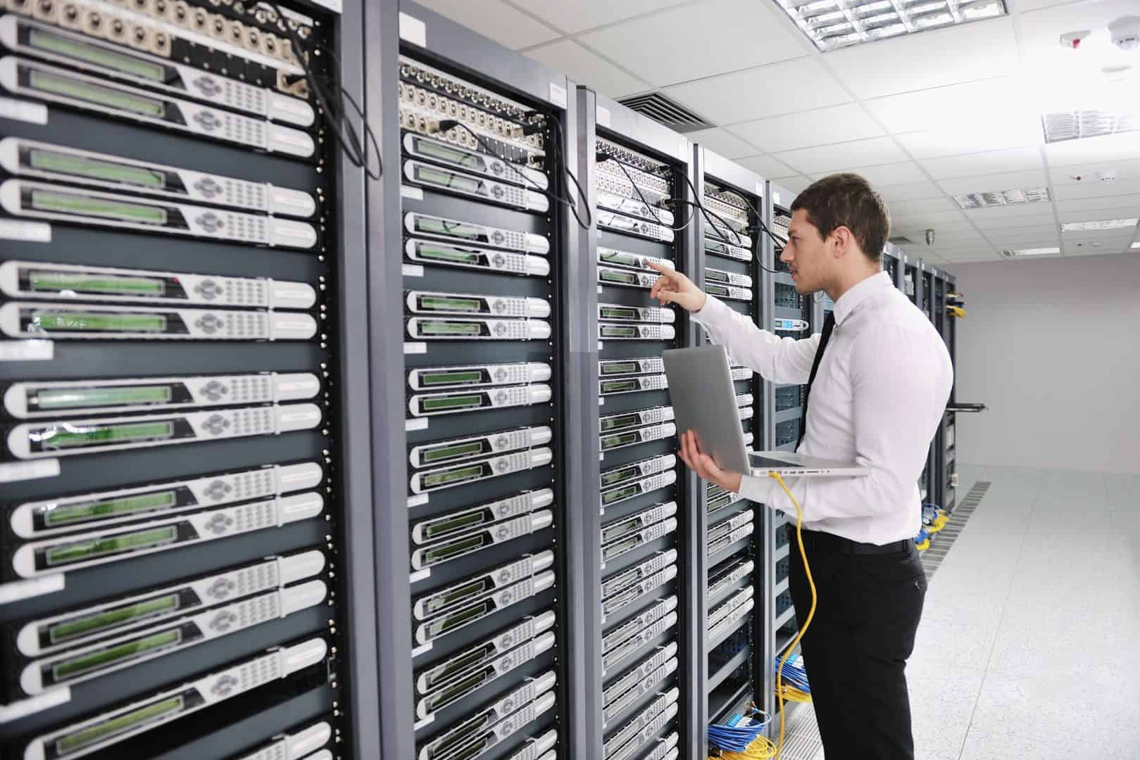 An IT professional stands in a server room inspecting network equipment, holding a laptop and pointing to a rack of servers, representing on-site IT support, infrastructure management, and data center operations.
