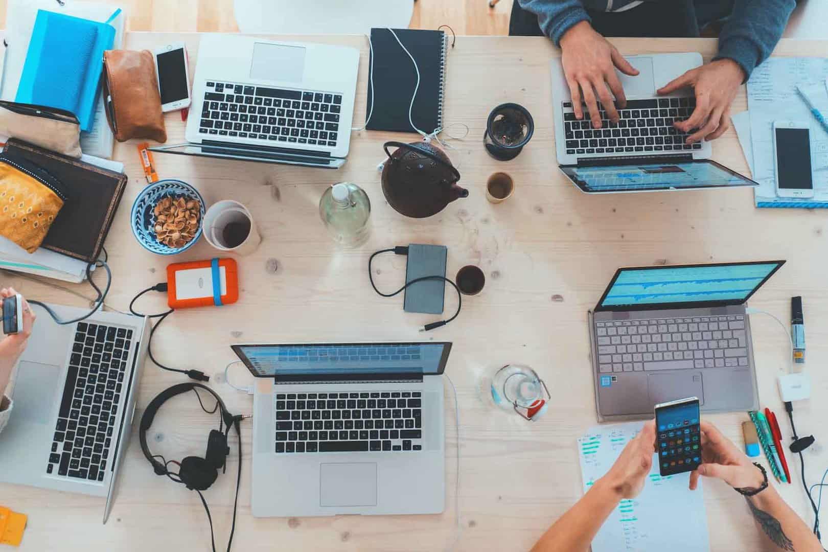 An overhead view of a shared workspace with several people working on laptops and mobile devices around a large table, with notebooks, coffee cups, and accessories visible, representing collaborative teamwork, digital workflows, and modern office productivity.