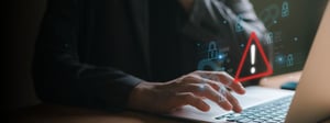Person using a laptop with digital security icons and a warning alert overlay, representing cybersecurity risks, threat detection, and proactive IT protection.