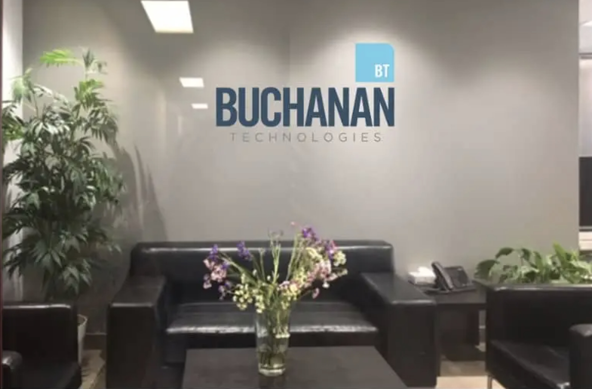 A modern office reception area with seating and plants, featuring the Buchanan Technologies logo displayed prominently on the wall, representing a professional corporate IT services environment.